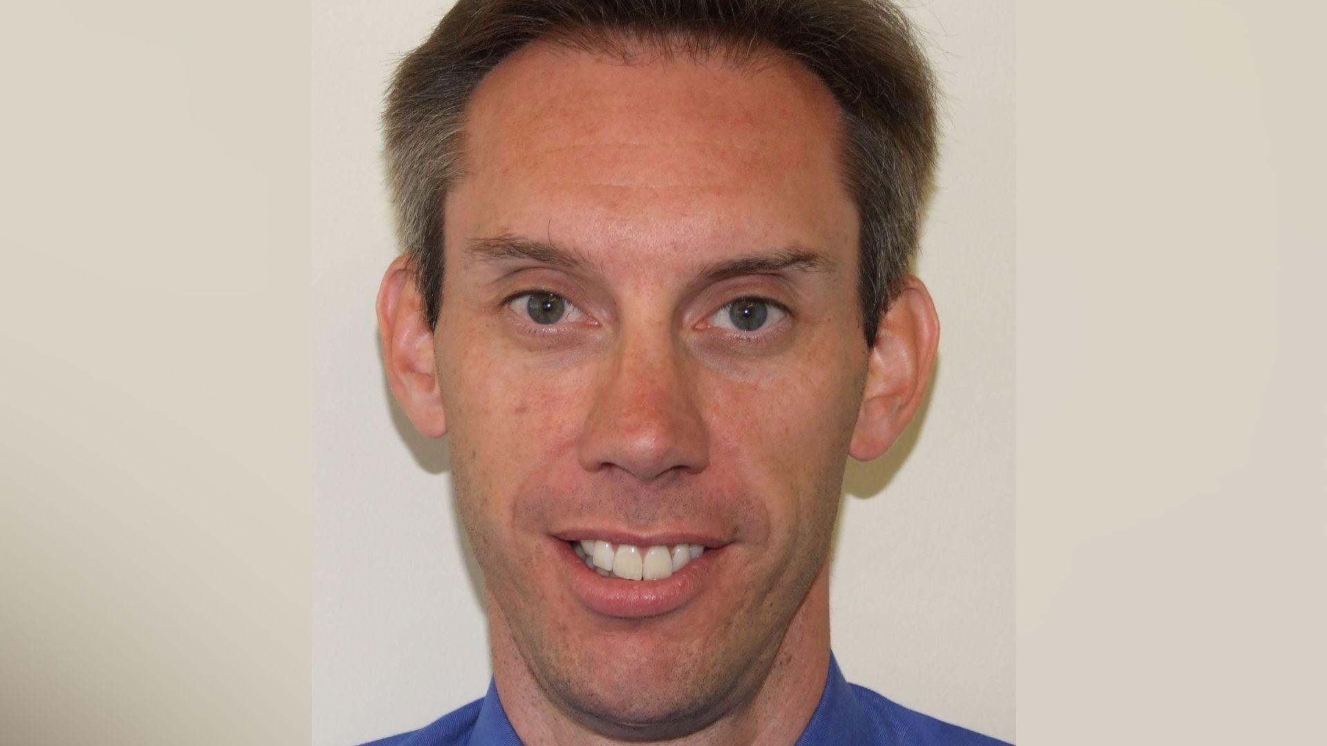 A man with dark hair, clean-shaven and smiling looking into the camera. He is wearing a blue shirt.