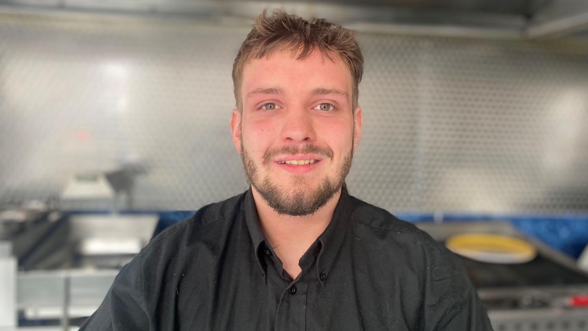 Alex is inside County Kebabs. He has light brown hair and beard and is wearing a black shirt.