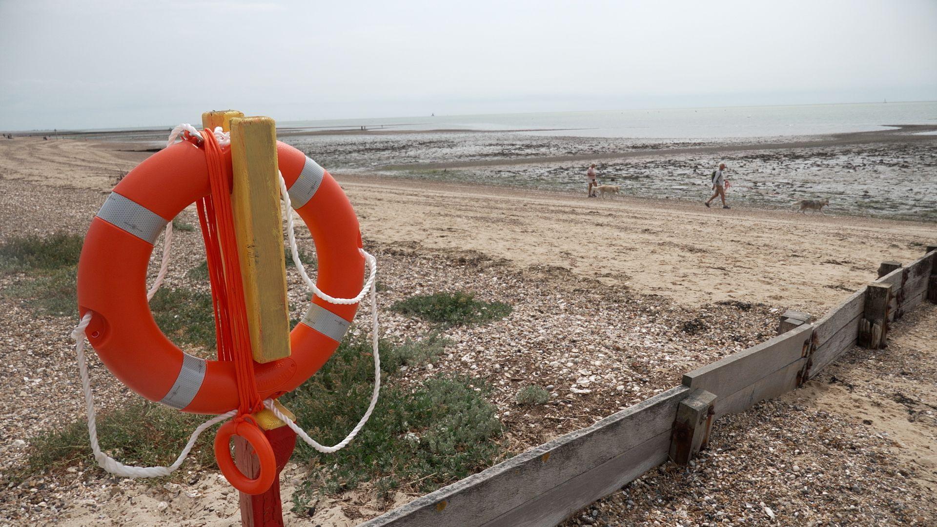A bright orange life ring hanging on a pillar. The life ring has two white stripes and has been propped up on the beach. Shingle, sand and the sea are in the background.