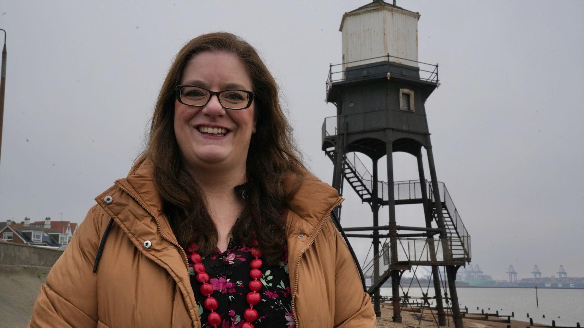 Jessica Bryan from Tendring District Council, standing in front of one of the Dovercourt lighthouses. She is wearing dark-rimmed glasses, a brown coat, a dark coloured floral top and a red beaded necklace.