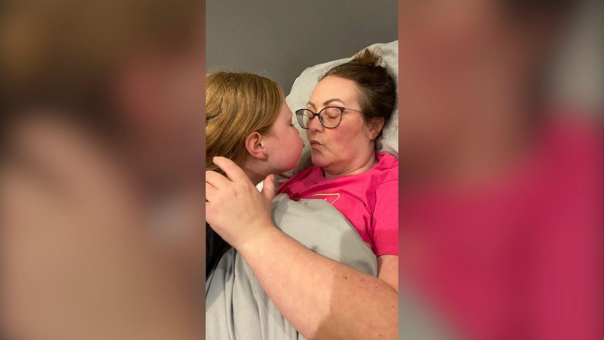 Michelle is lying in a bed, wearing a pink t-shirt and glasses. Her daughter Connie is leaning over her mum to give her a kiss