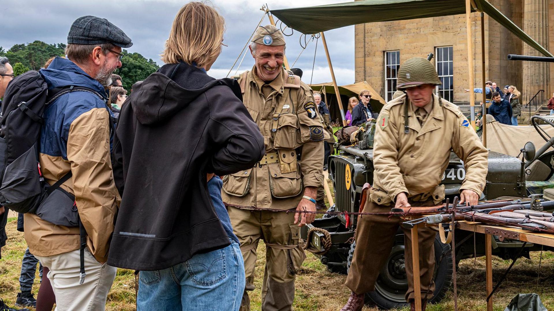 Two men in Second World War uniforms stand next to a table full of ammunition, talking to a couple of visitors with Belsay Hall behind.