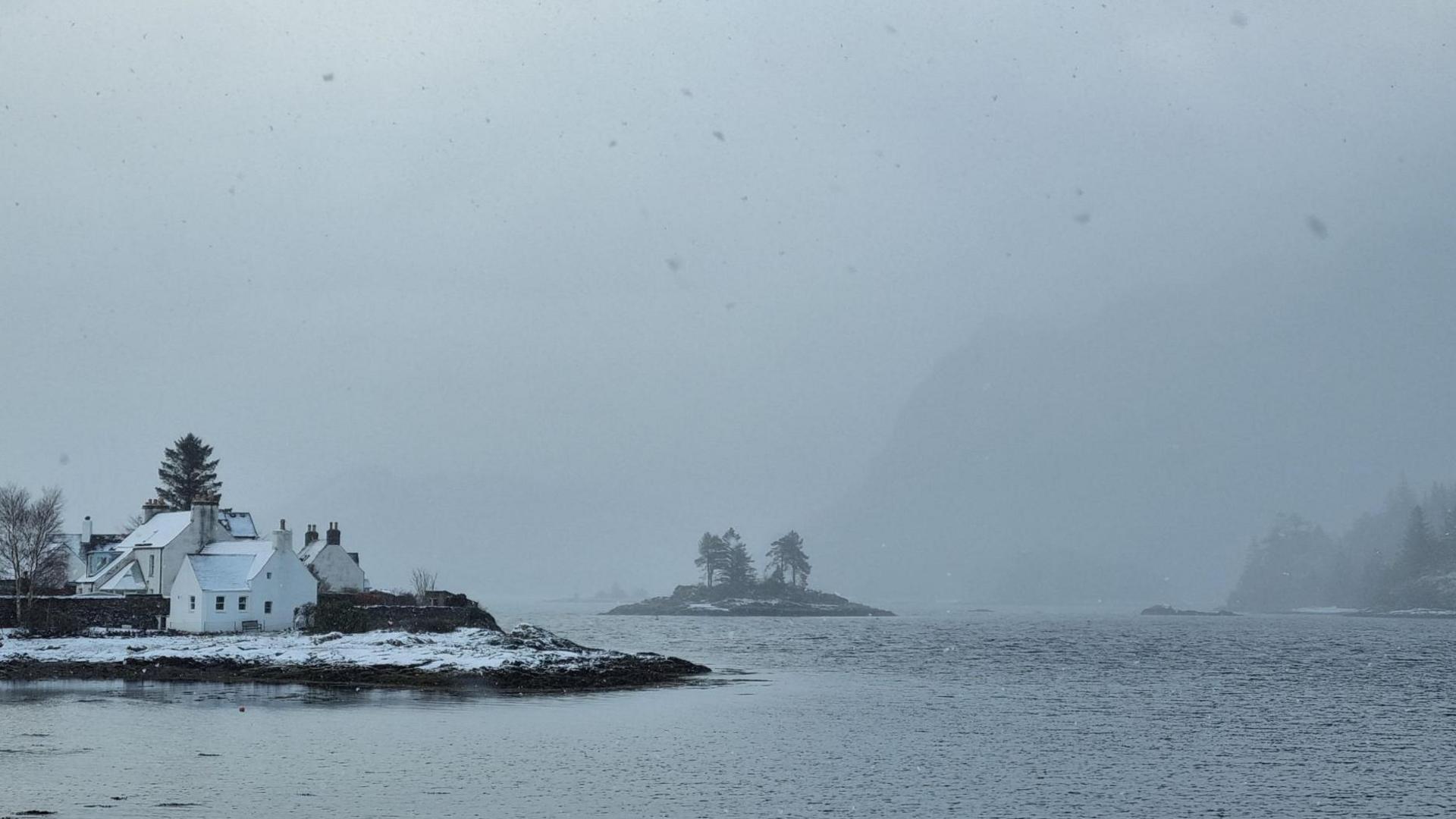 White-walled houses on a shoreline in the Highland village. There is a small island with trees on it. A heavy snow shower obscures hills in the distance.