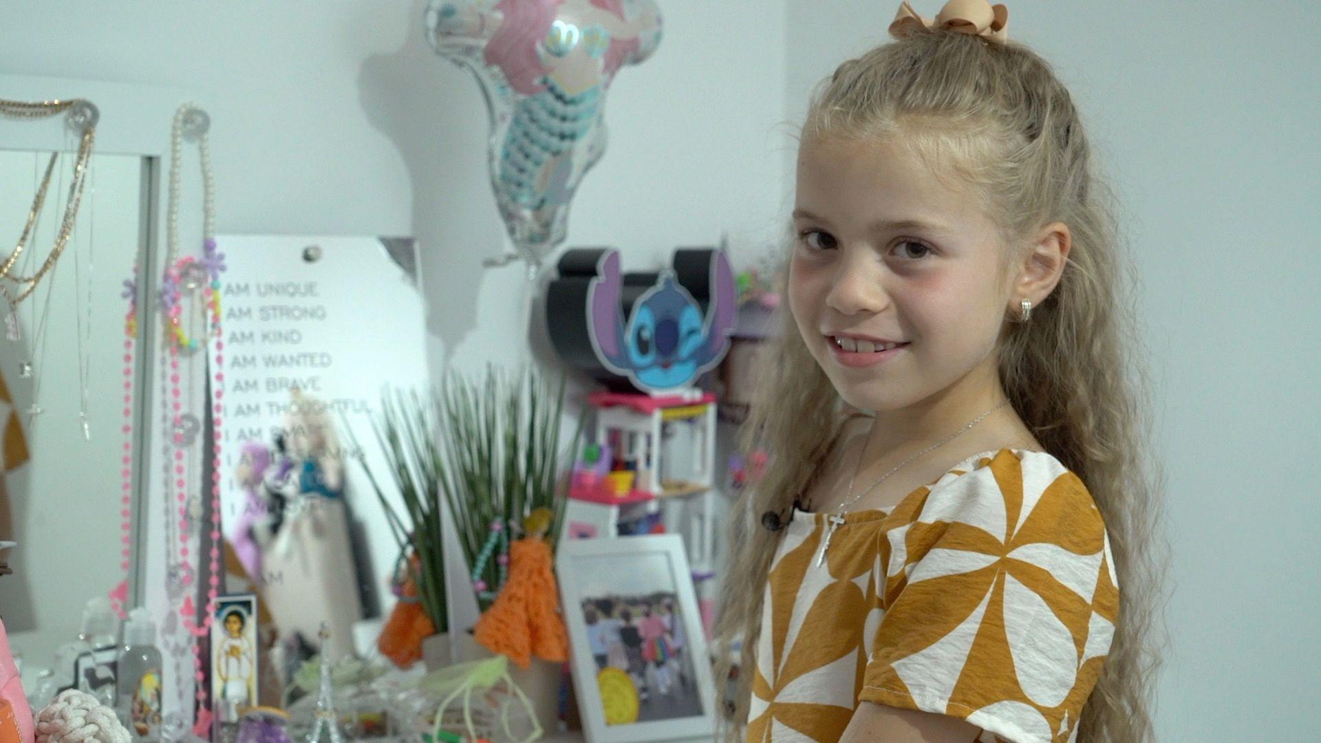 A young girl stands in front of of a selection of toys, jewellery and a balloon. She has blonde hair which is half tied back off her face. She is wearing a mustard and white-patterned dress.
