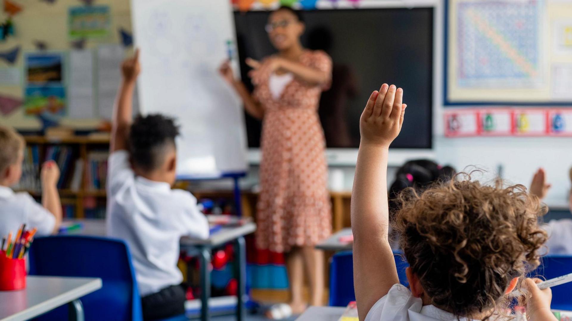 A teacher in a classroom standing at the front of a class as two children hold their arms up in the air. The teacher is wearing a dress and pointing, the children have their backs to the camera 