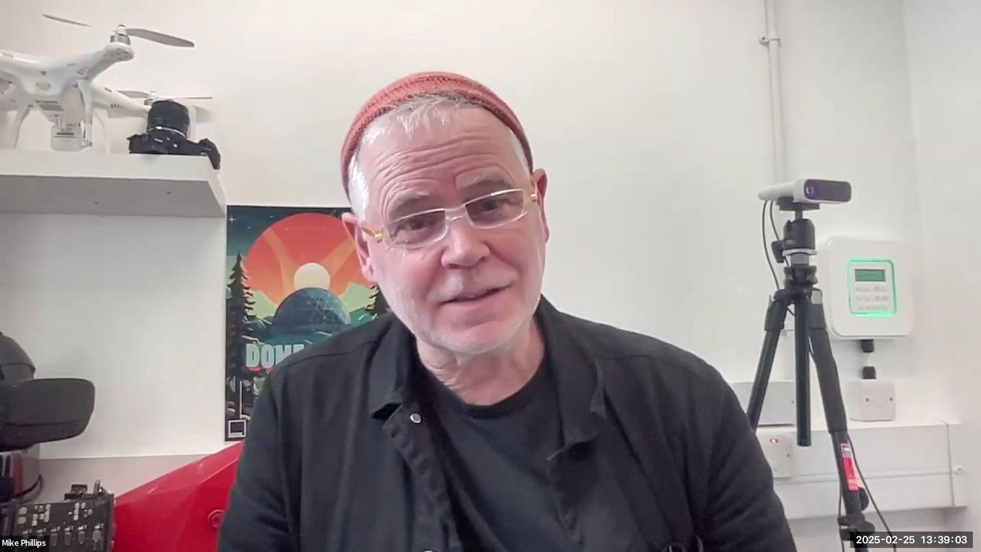 A man with an orange hat and black shirt sits beside drones and a camera on a tripod.