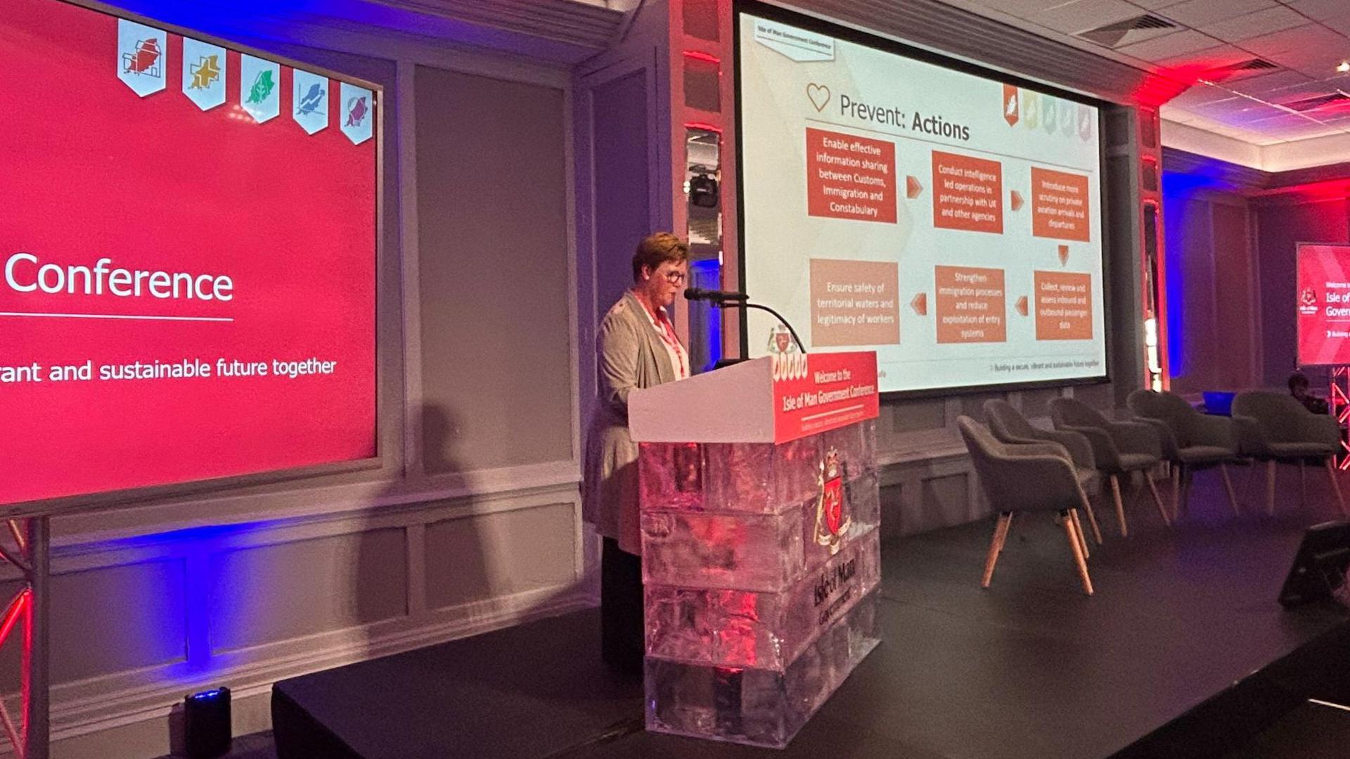 Jane Poole-Wilson standing at a lectern speaking into a microphone. She is wearing a beige cardigan and has short auburn hair and is in front of a large screen with other large pink screens nearby. 