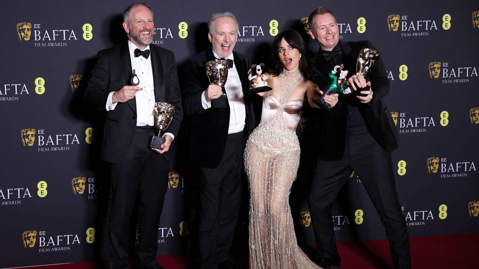 Three men stand on the red carpet holding figurines of Wallace and Gromit. They are dressed in suits, for an awards ceremony. They are joined by US singer Camilla Cabello who presented the award. She is wearing a rose gold dress with silver detailing. 