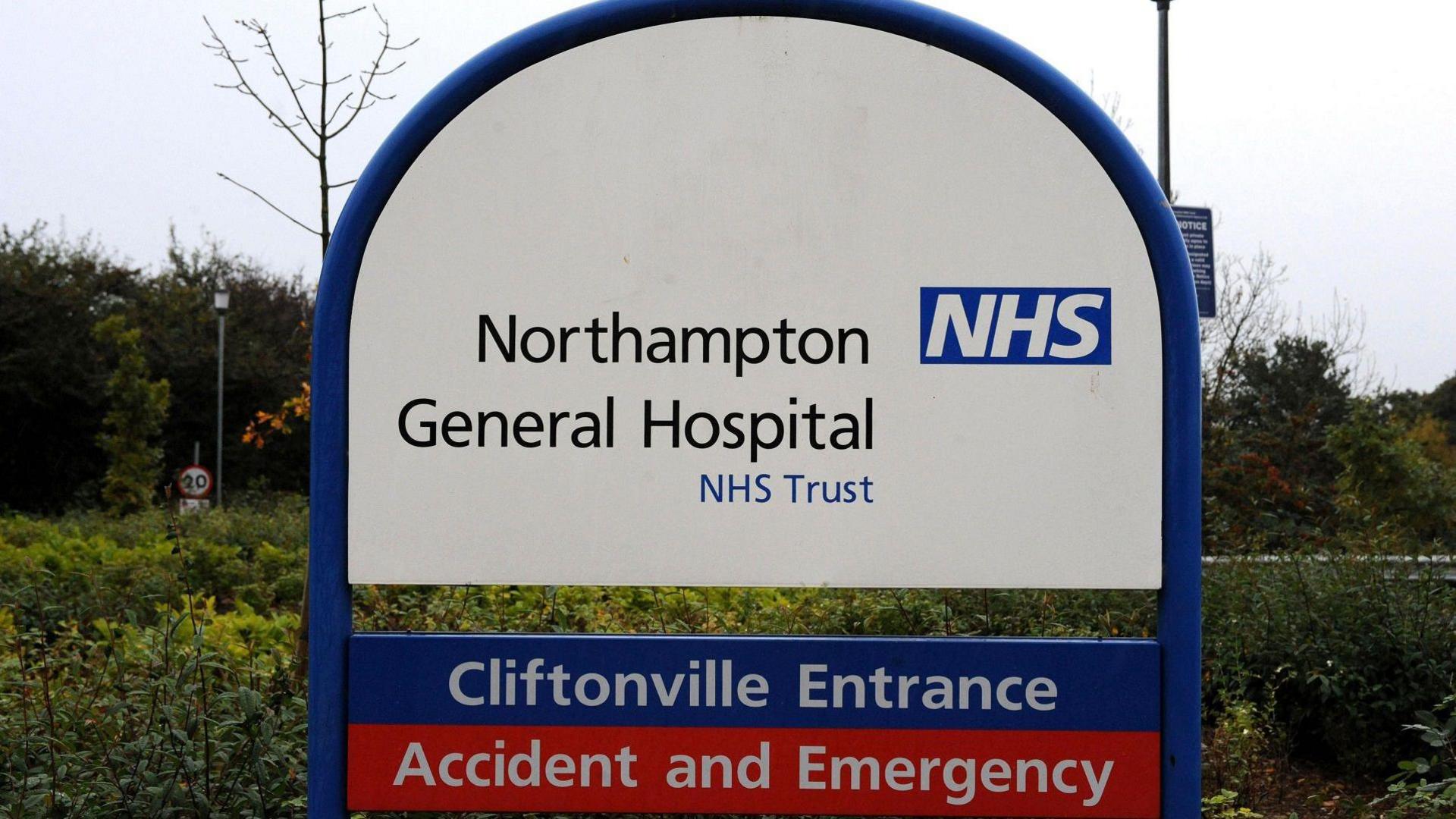 A white and blue sign next to bushes at the hospital. It says Northampton General Hospital NHS Trust, then below: Cliftonville Entrance, and in a red strip below that: Accident and Emergency.