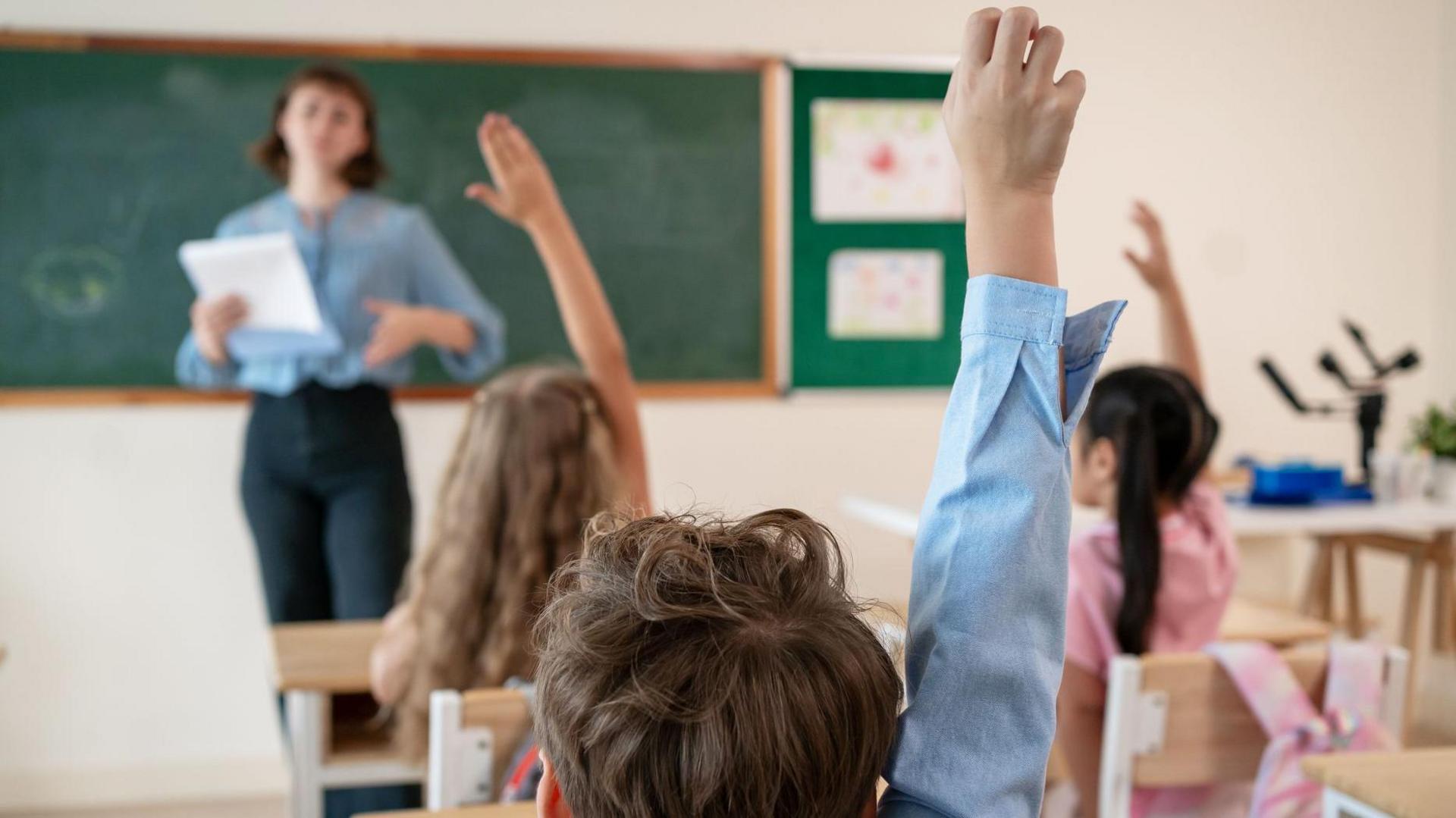 A classroom with three students sitting on chairs and a teacher at the front who is out of focus. The teacher has short brown hair and is wearing a blue shirt and black trousers. The children have a pink and blue shirt on, one has long brown hair, the other black piggy tails and the boy short brown hair. They are raising their hands.