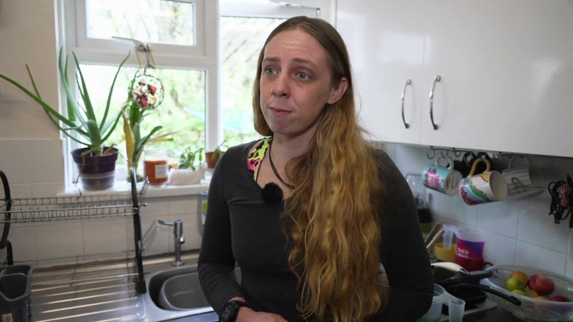 Morwenna Roesner stood in her kitchen. She is looking to the right and is stood in front of a white cupboard and window to the right. She is wearing a black top and has long hair.