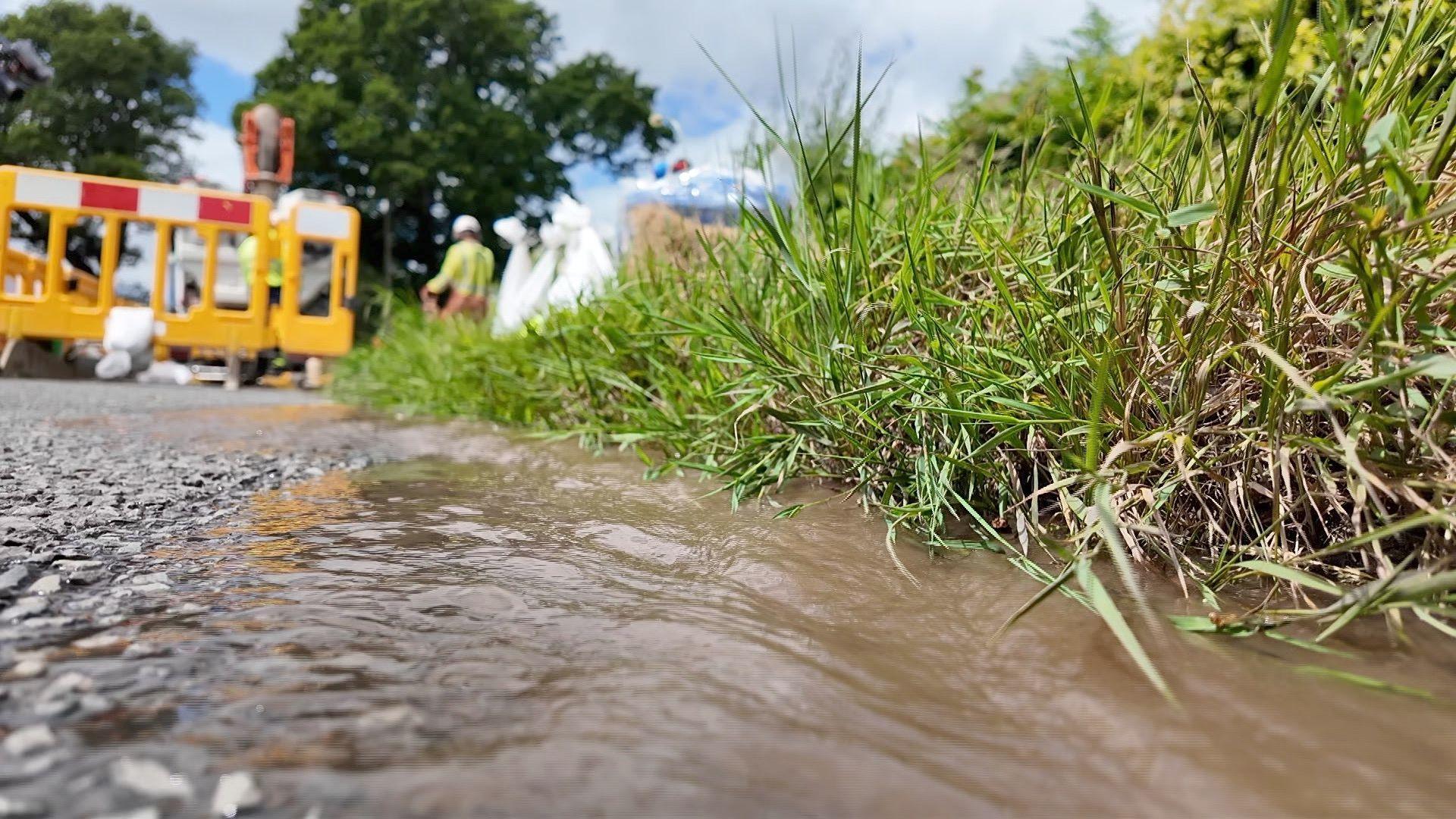 An image of a road in the countryside with water running down the edge of the carriageway. An area of road is fenced off with people in high viz clothing working to fix a leak.