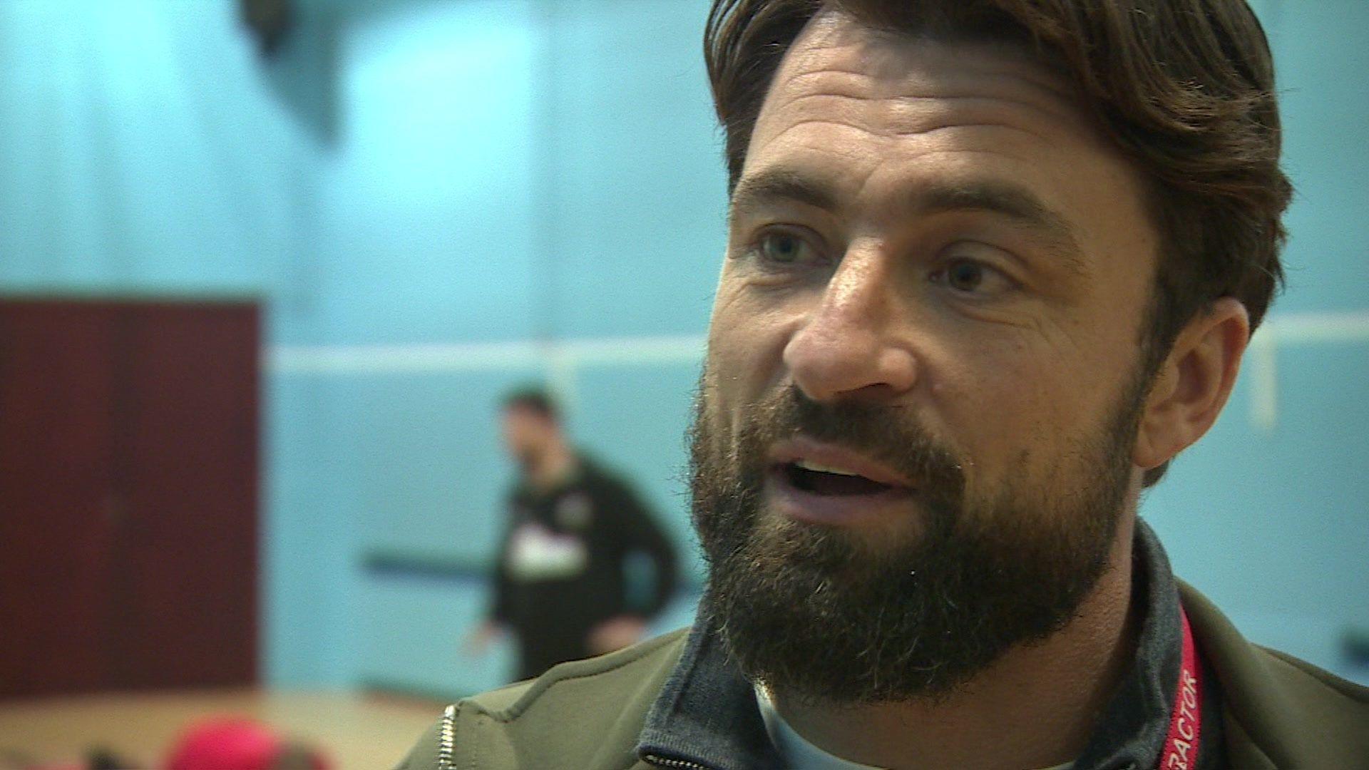 A close up photo of Russell Martin, with dark hair and a beard, wearing a green jacket. He is standing in a gym with light blue walls, with a blurred figure of two men playing sport behind him.