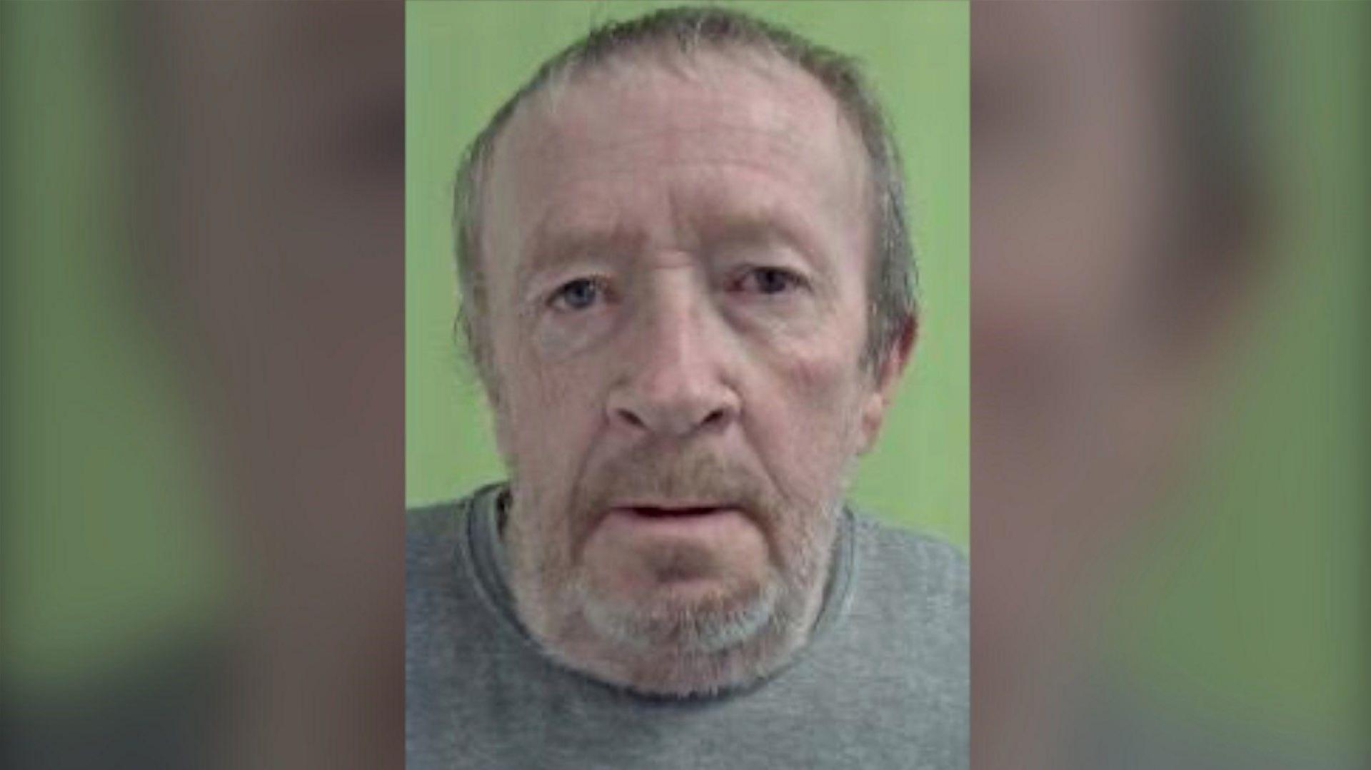 A police mugshot of a man in his 60s wearing a grey crew-necked shirt. He has short, greying brown hair and a short beard. 