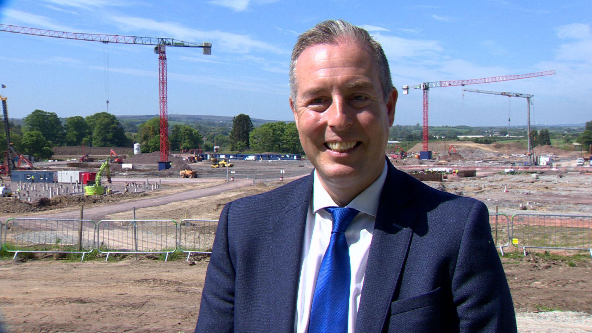Paul Givan. He has grey-ish hair, wearing a blue blazer, white shirt and blue tie. A construction site is behind him.