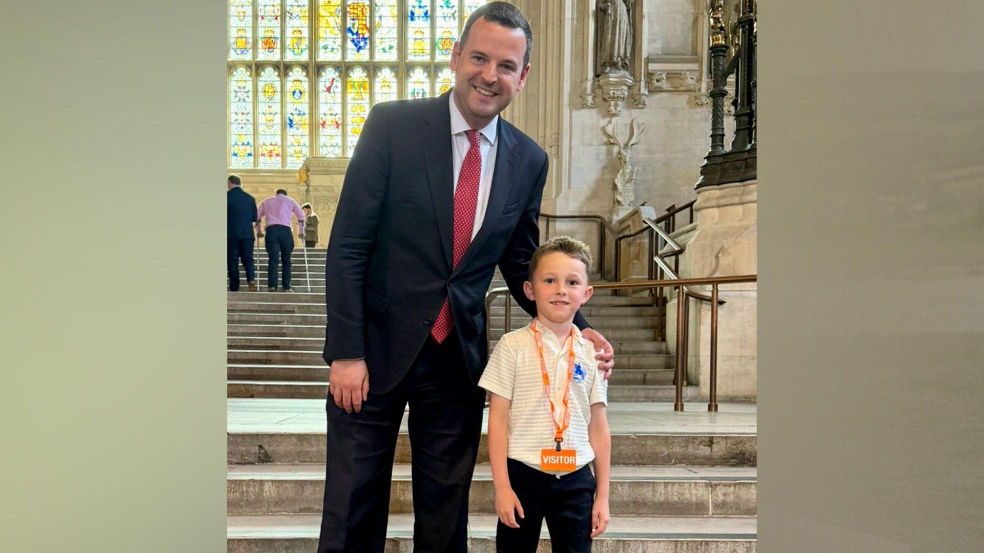 Teddy standing on a stone staircase with a stained glass window behind. The six-year-old boy is wearing a yellow shirt with an orange lanyard around his neck. He is standing with MP Paul Holmes who has his arm around Teddy's shoulder. He is wearing a dark suit with a red tie.