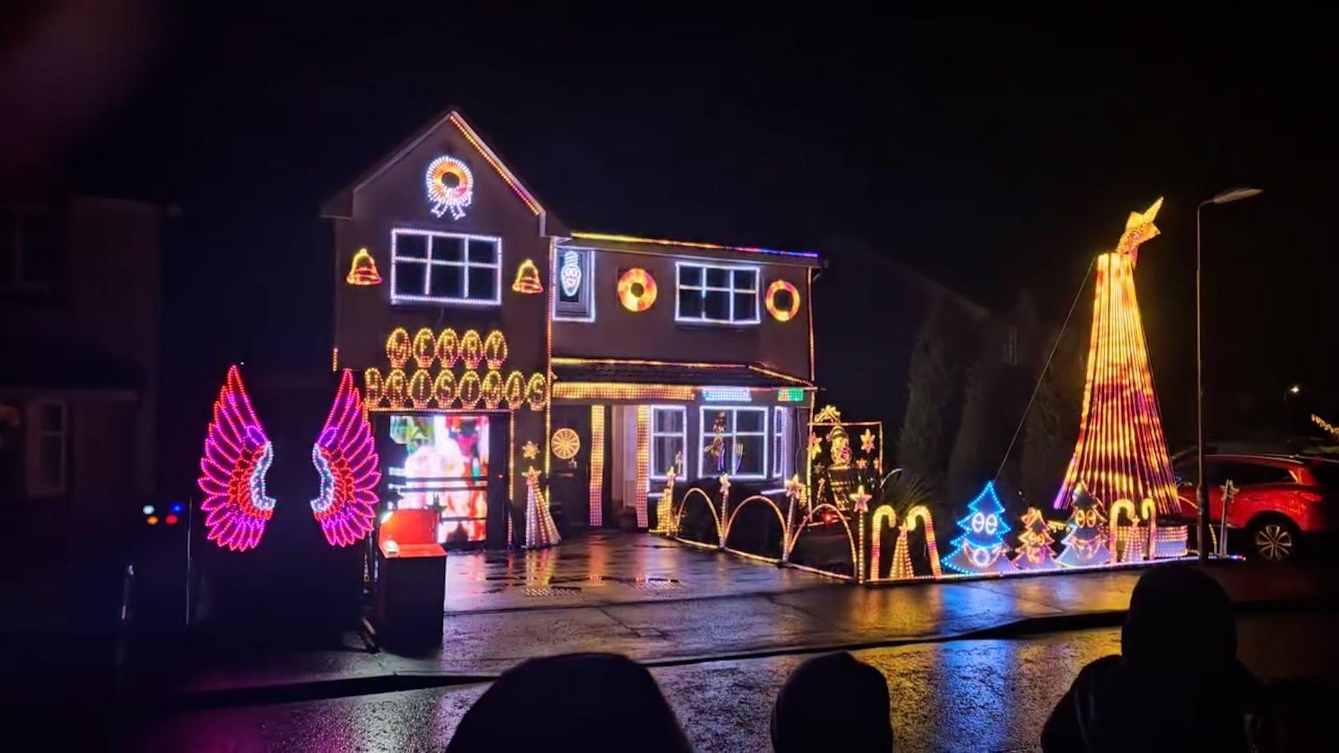 A two-storey house brightly illuminated in multi-coloured Christmas lights. There are small light up Christmas trees, light up candy canes, pink light up angels wings around the garden.