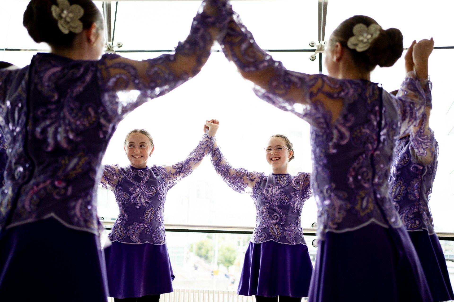 A group of céilí dancers, all wearing matching purple dresses. They are holding hands aloft in a circle