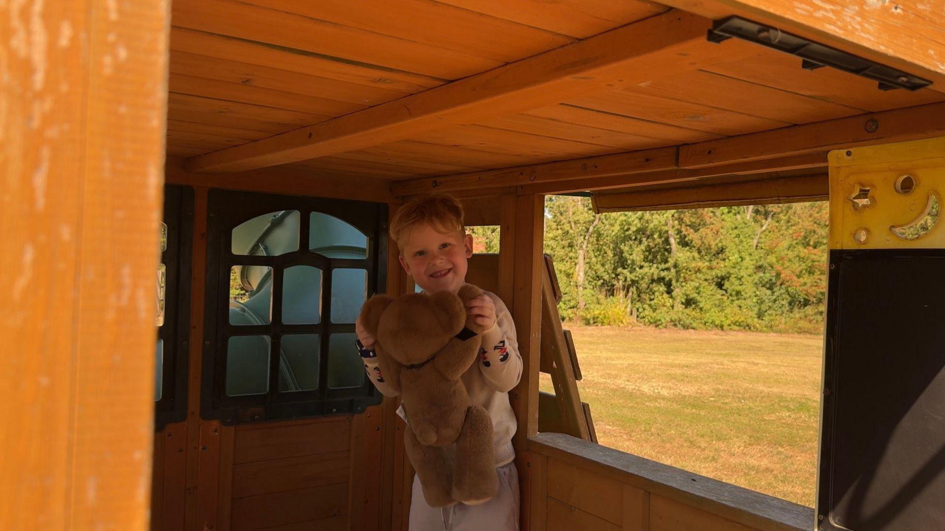 Monty is a young boy with blonde hair. He is wearing a beige tracksuit and is stood holding a large teddy bear in a wooden playhouse in a garden. He is holding it by both arms and smiling. We can see grass and trees in the background through the window if the playhouse.