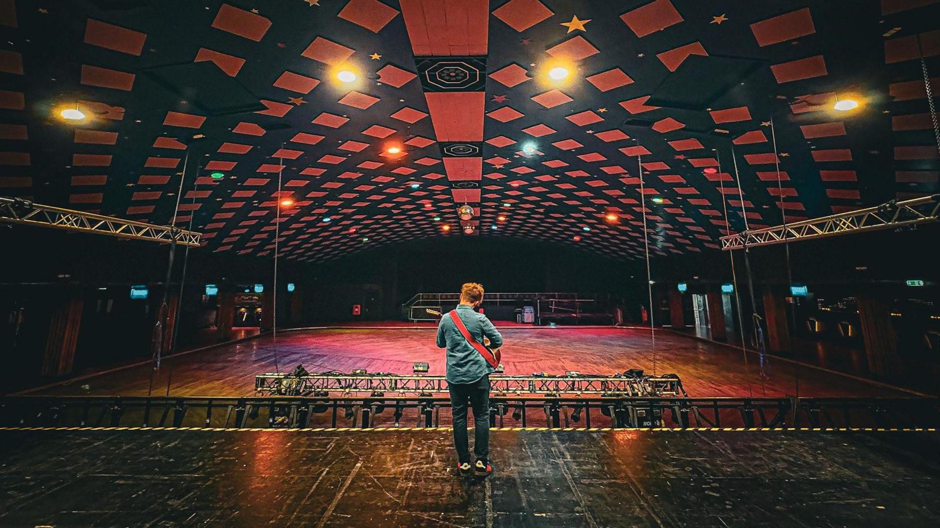 A man playing a guitar standing on the middle of a stage looking on to an empty auditorium