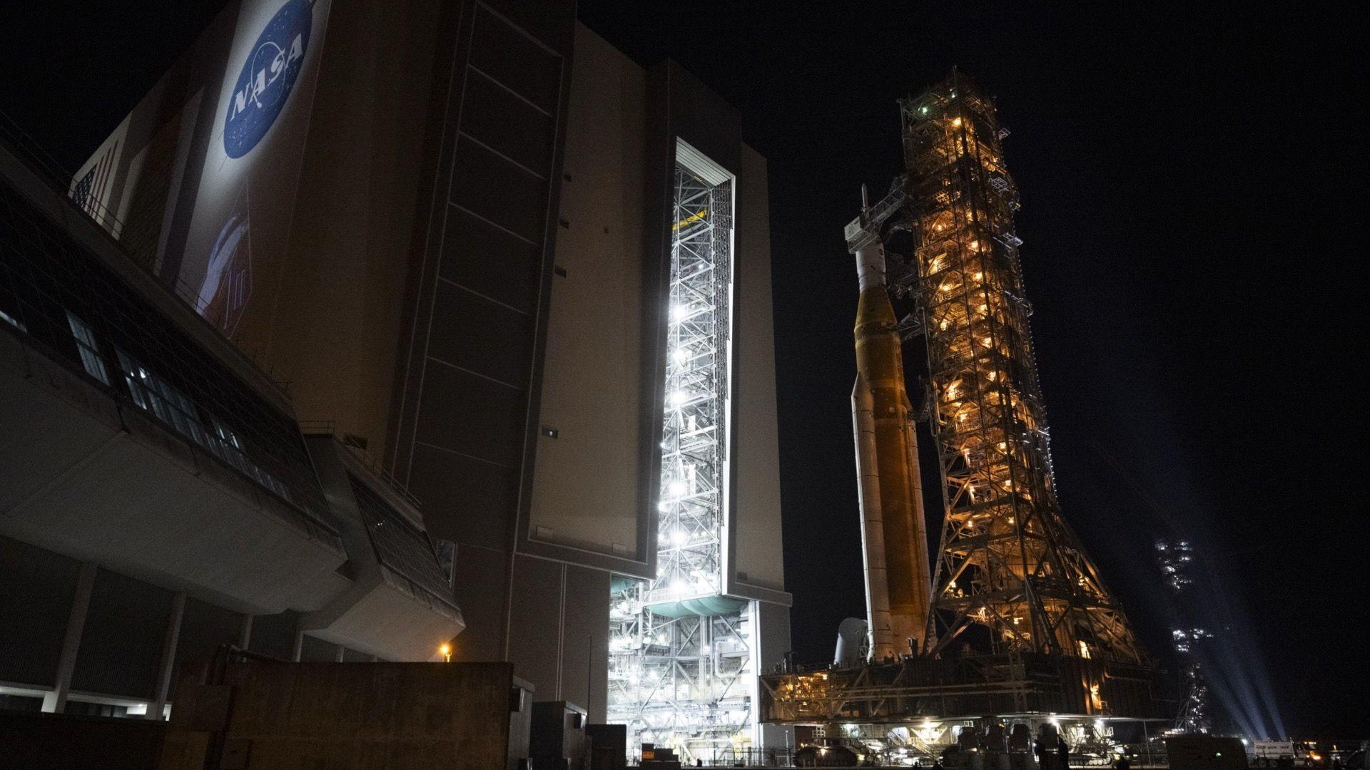A large rocket, secured to a mobile launcher, getting rolled out of a Nasa hangar whilst it's dark outside.