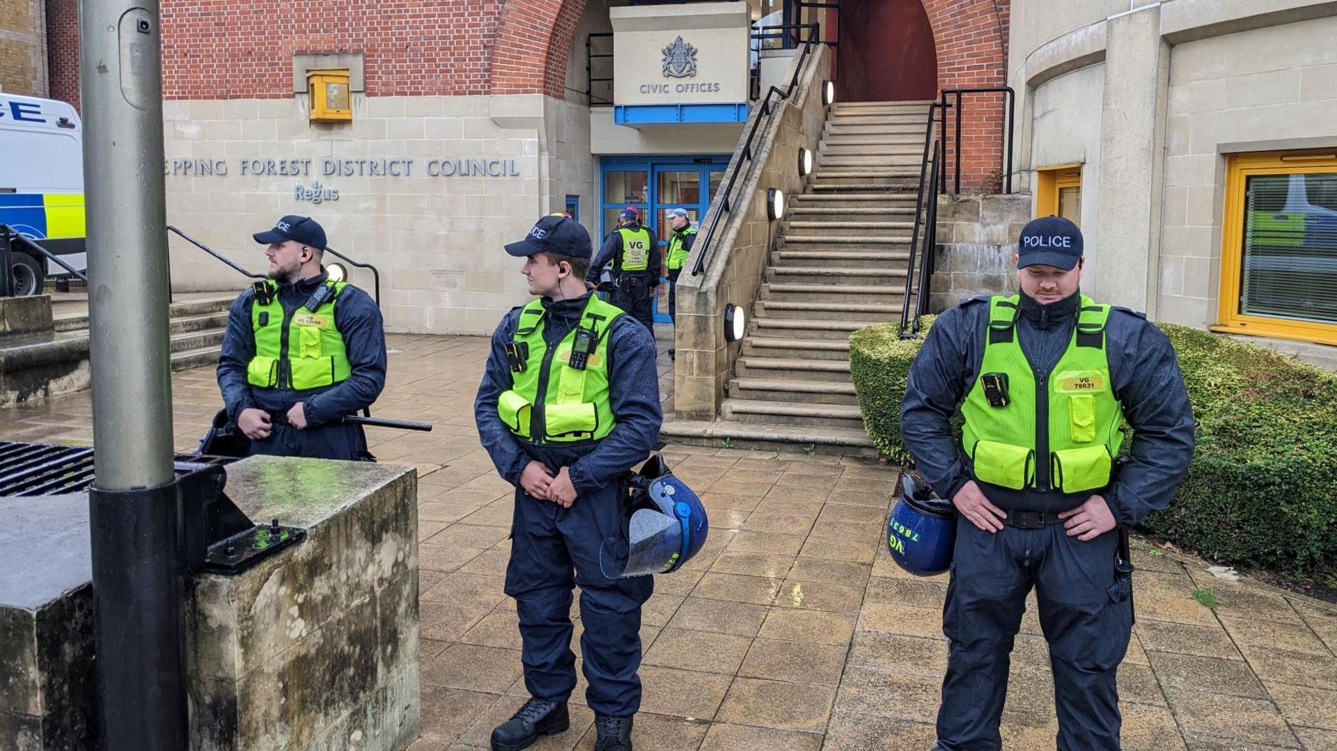 Three police officers in thick jackets and high-vis vests standing in the foreground outside the Epping Forest District Council building.