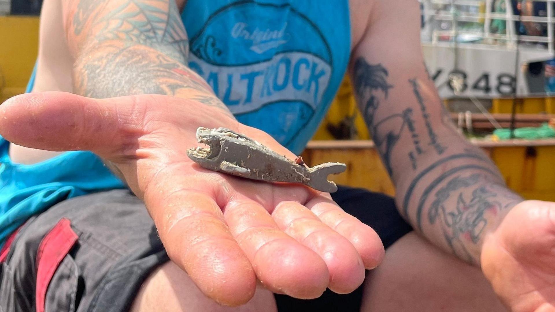 Richard West is sitting on a trawler and has stretched his open palm out towards the camera. In his hand is a pale grey barnacle-covered Lego shark and he is smiling. He has tattoos on his arms and is wearing a blue cutaway Saltrock T-shirt and a backwards cap.