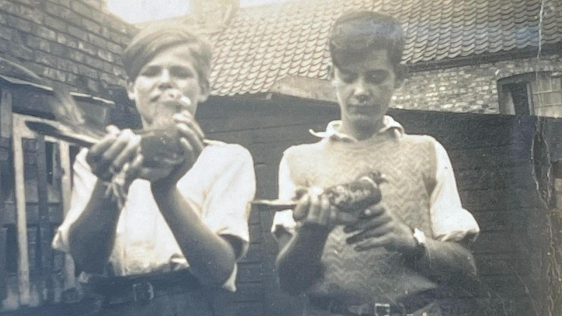 The image is a black and white picture of two boys both holding pigeons