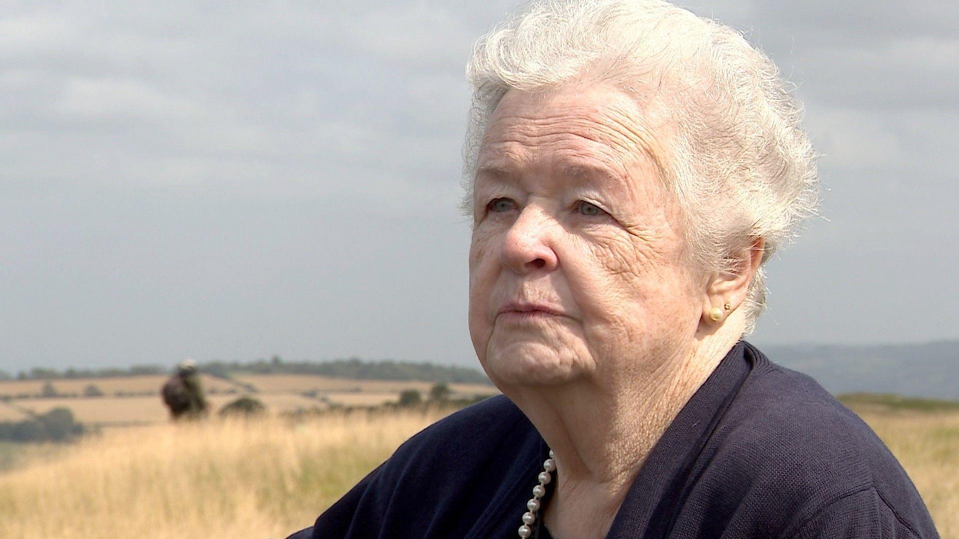 Maureen Sullivan, with white curly hair wearing a blue jumper and pearls, on a hilltop, looking off-camera