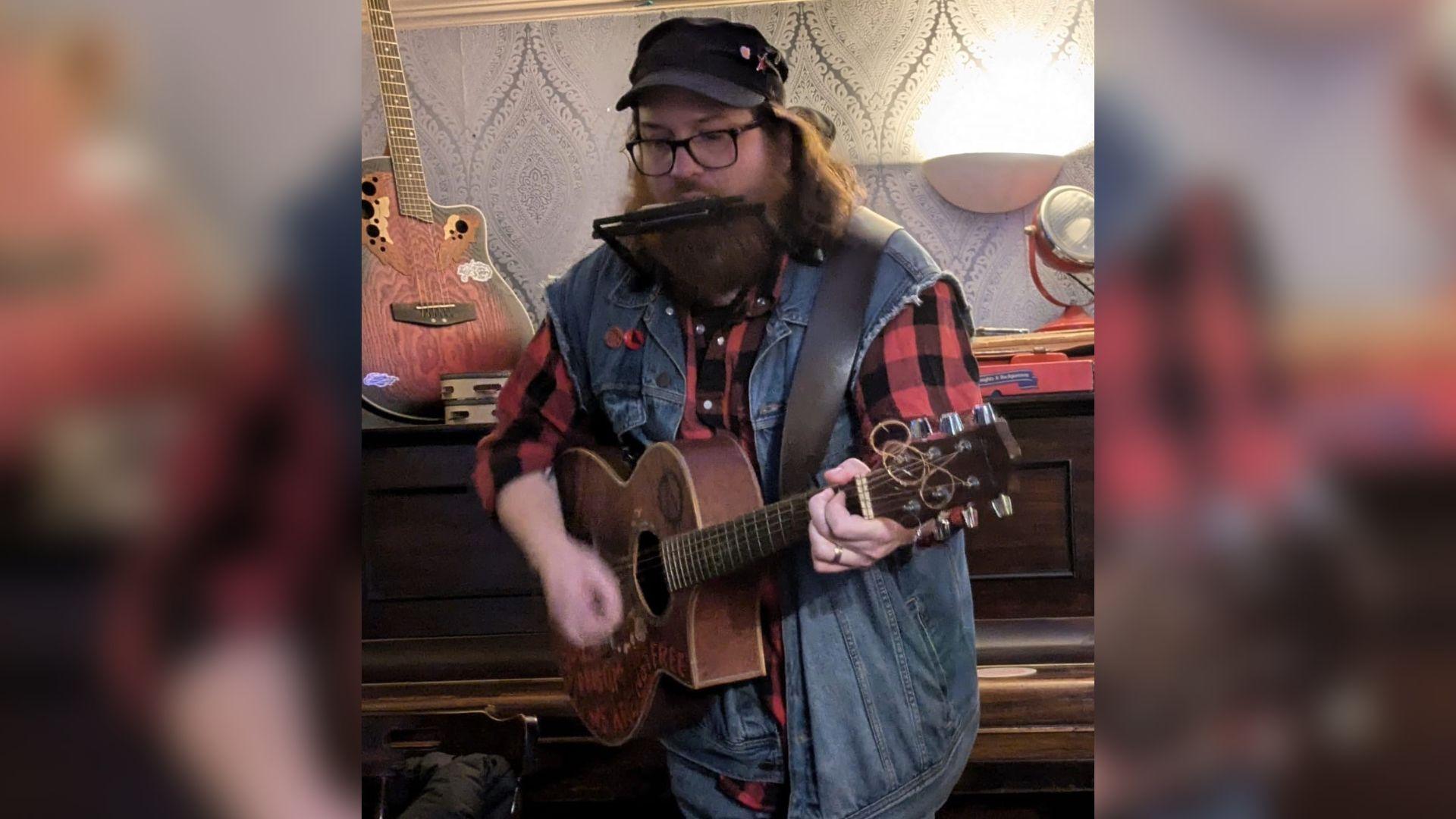 Llun o Joshua Gardner yn perfformio gan chwarae'r gitar ac harmonica ar yr un pryd.