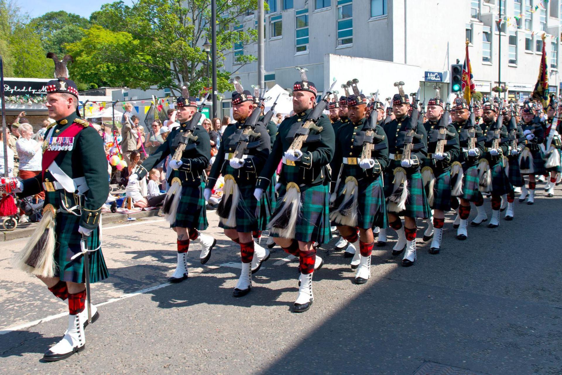 Soldiers from The Royal Regiment of Scotland marching along a street in full Highland dress, including kilts, and carrying weapons on their shoulders.