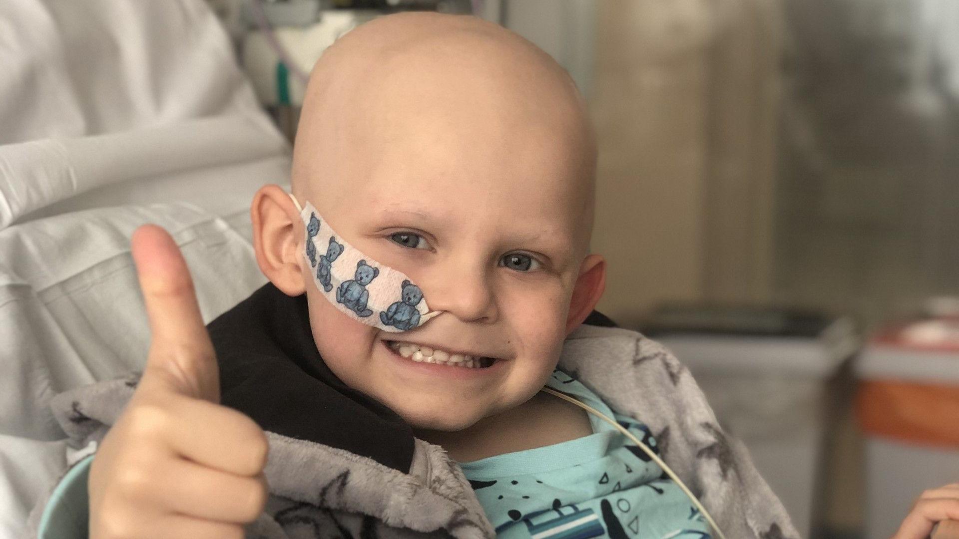 A boy in a hospital bed smiling and giving a thumbs up. He has no hair and a plaster with blue teddies securing a drip line to his right cheek. He is wearing a grey dressing gown with stars on it and turquoise pyjamas.