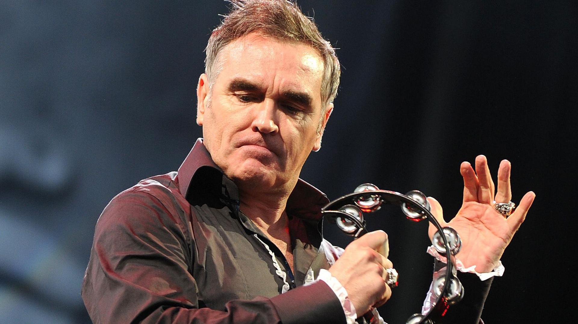 Morrissey, wearing a black open-necked shirt, bangs a black tambourine on his outstretched hand