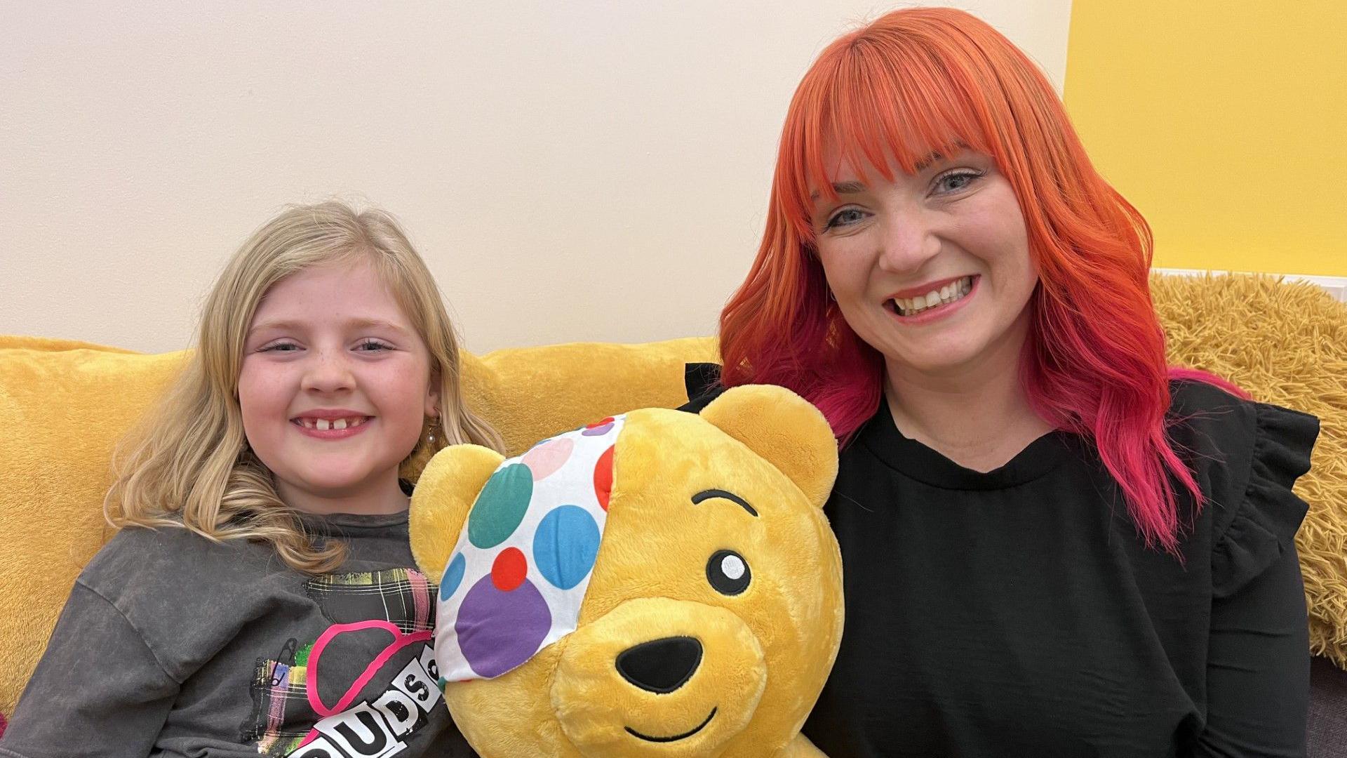 A young girl, with blonde hair and blue eyes, wearing a grey t-shirt with 'Pudsey' on it, is sat on a yellow sofa next to an older women who has bright orange and pink hair, with a box fringe. She is wearing a black top. In between them is a toy Pudsey bear. Both are smiling directly at the camera.