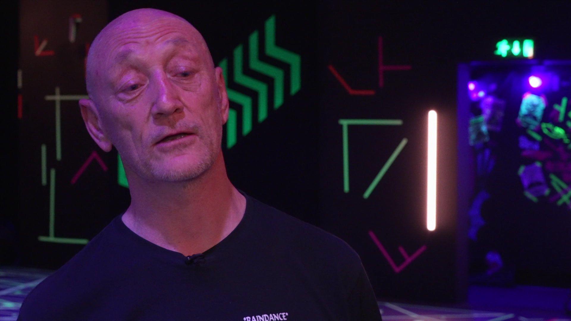 A hairless middle-aged man on a dancefloor with fluorescent lights behind him