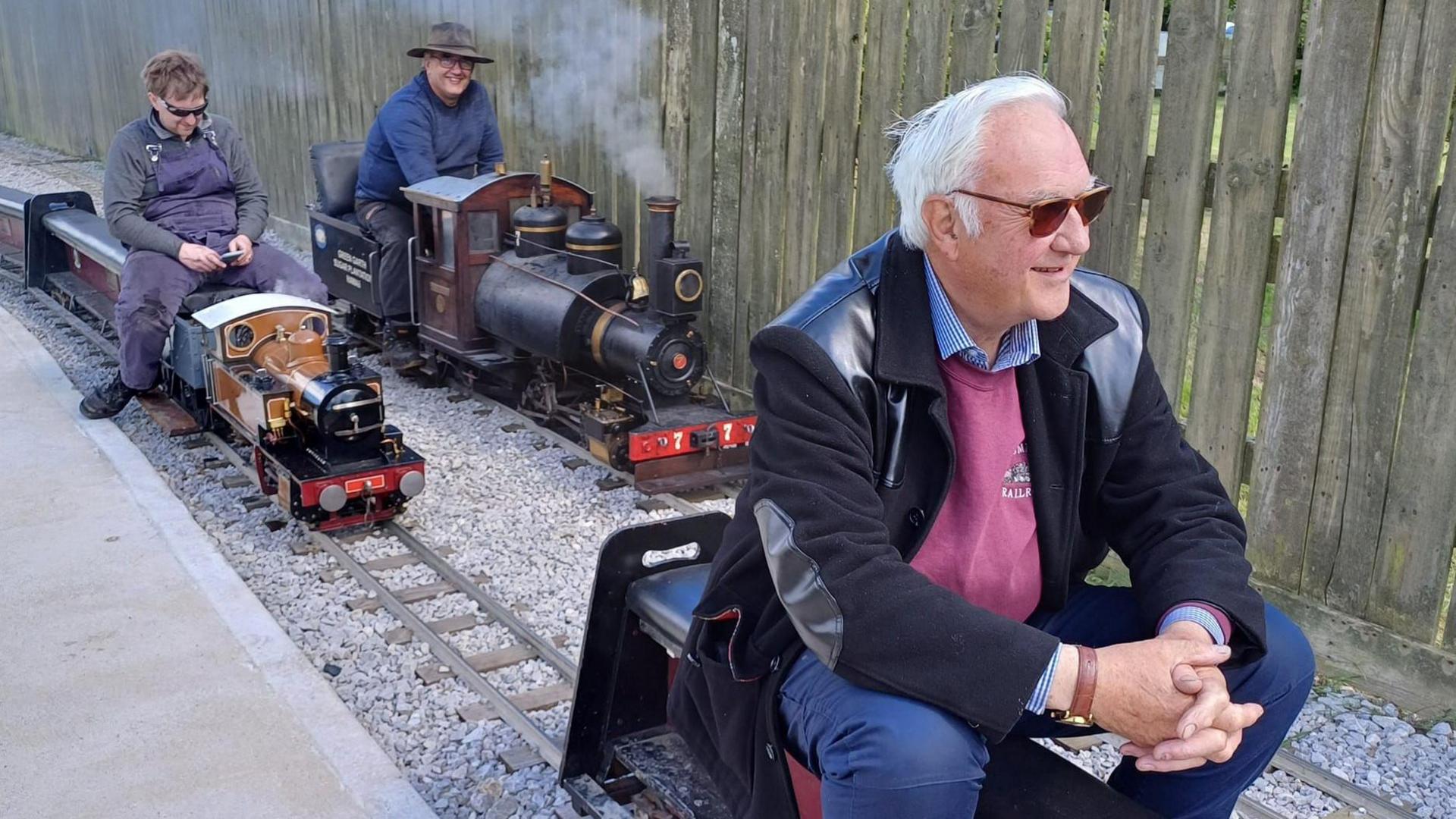 Three men sit on three miniature trains on two rail tracks. The man at the front has white hair, sunglasses, a pink jumper and black jacket- he has his hands folded in front of him and looks away. The man back left has blue overalls, black sunglasses and looks down at his phone. The final man on the right smiles at the camera - he has a wide brimmed hat and a blue jumper on.