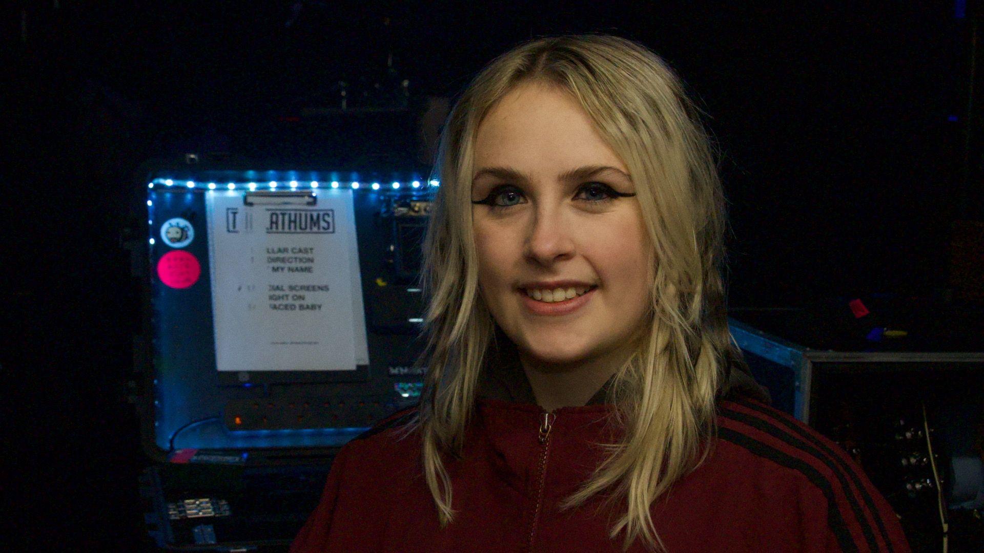 Bridget has blonde shoulder-length hair. She wears a dark red tracksuit top which is zipped up. She has dark eyeliner and is smiling at the camera. She stands in front of a music mixing desk.