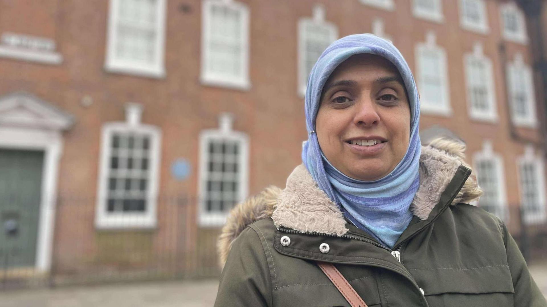 A woman in a coat and head scarf stood in front of a grand red brick building