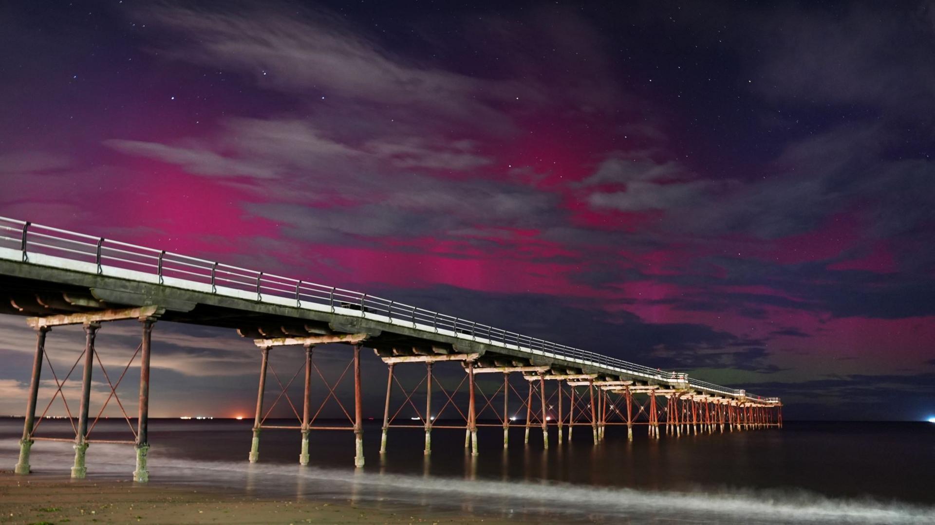 A pier stretching out across the water at night time on tall metal legs. The sky glows purple and pink, partially obscured by grey clouds dotted around.