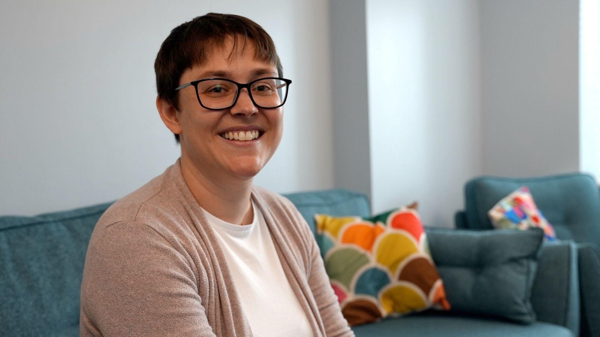 Emma is sat on a teal-coloured settee, with a brightly coloured cushion in the background. She has short brown hair and is wearing dark-framed glasses. She has a white top and beige cardigan on.