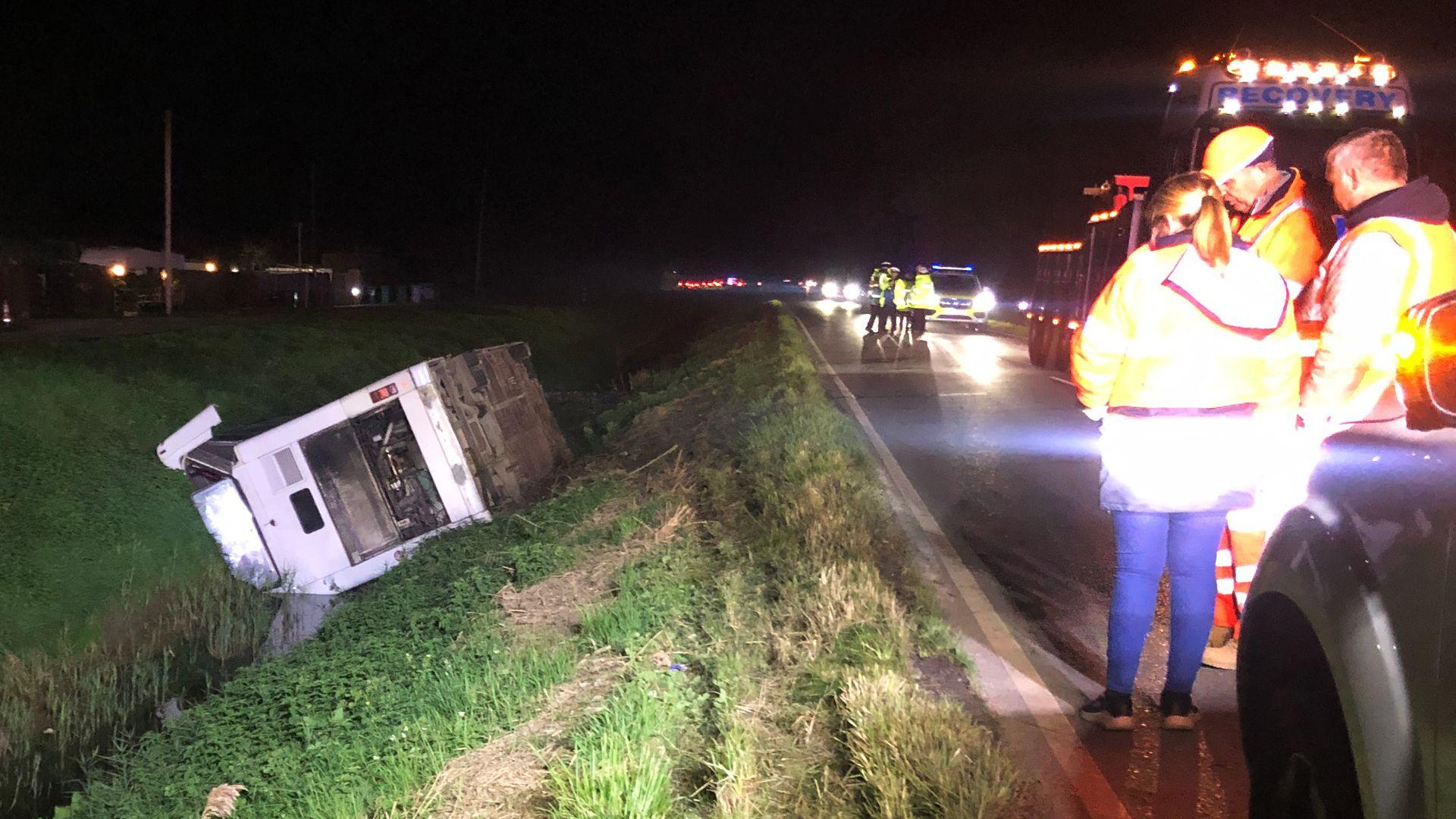 The bus on its side in a ditch. In the foreground are people in hi-vis jackets and a recovery van.