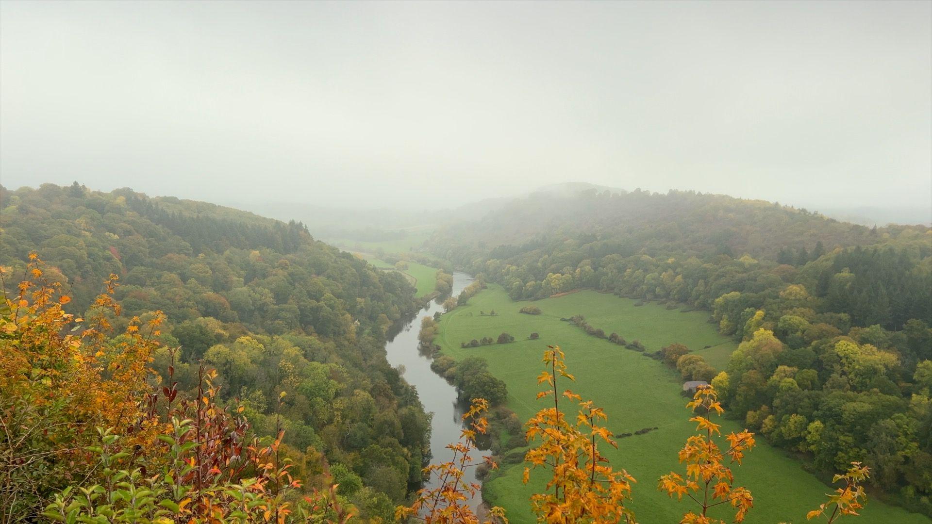 A foggy day in the Forest of Dean. It's a beautiful countryside scene with Forest on the left hand side of the photo. In the middle a river winds it's way into the distance whilst green fields sit on the right hand side with more forest sitting further across. The sky is grey and white obscuring the view in the distance.