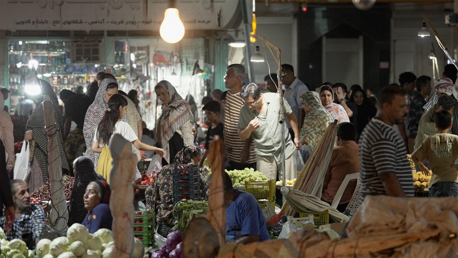 A busy market scene with lots of people looking and stalls selling vegetables. The stall front left appears to be piled with white cabbages and a man and a woman are sitting behind them. 