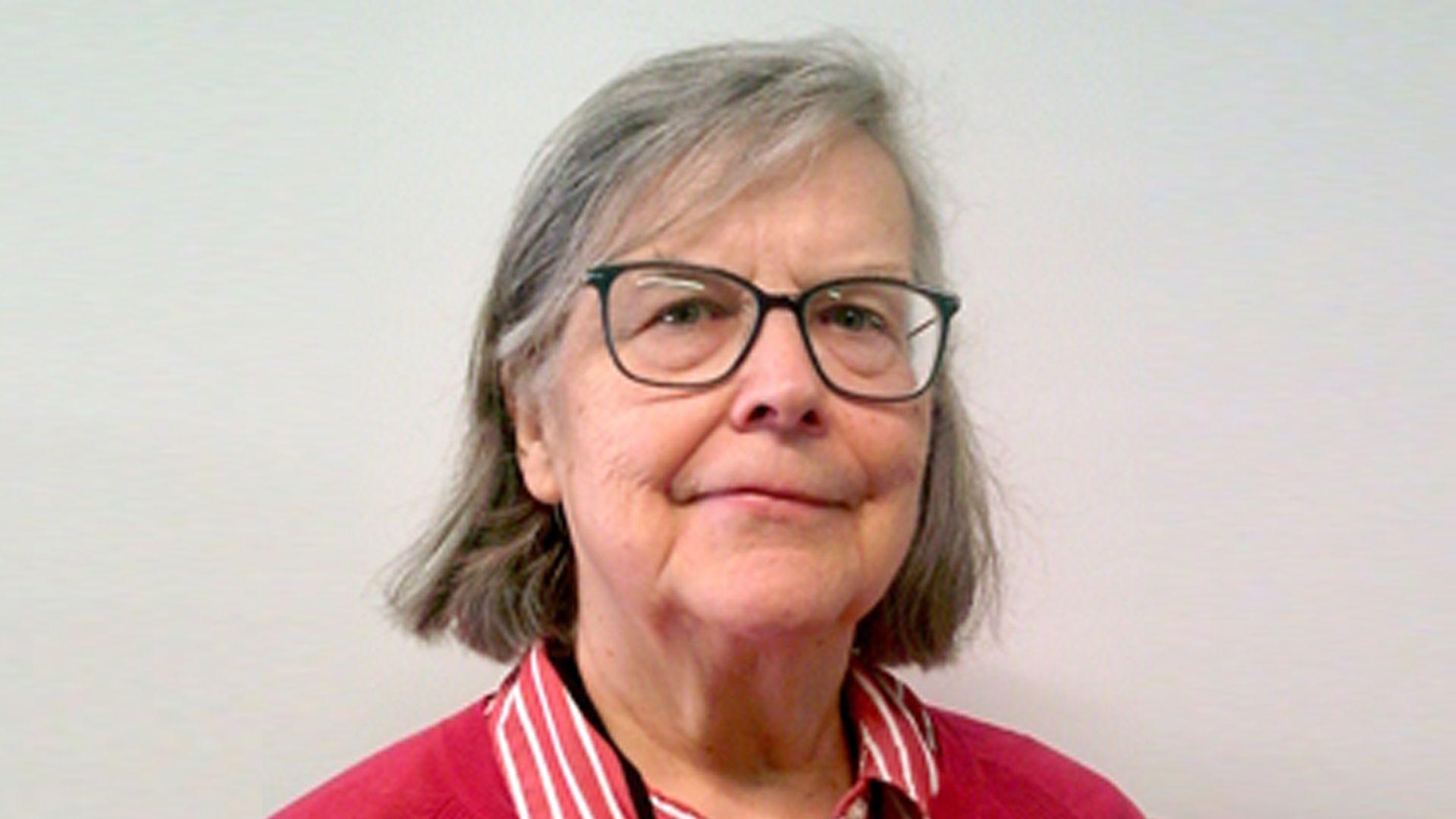 A headshot of a woman with long grey hair, glasses and wearing a red top.