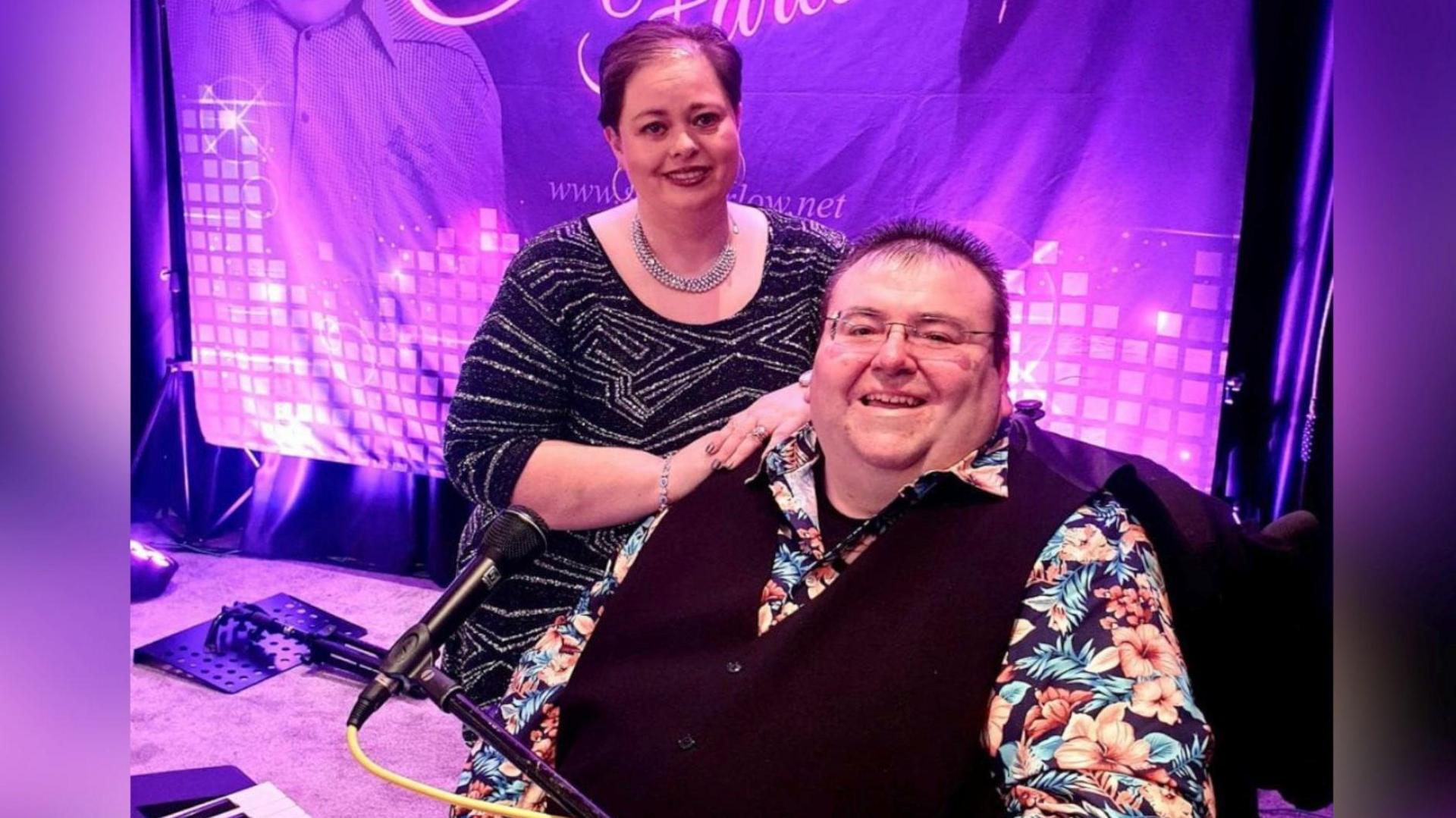 Stephen sits, while his wife Pauline stands and rests her hands on his shoulder. Stephen has brown hair which is slightly spikey, glasses and wears a bright hawaaian shirt, with a black waistcoat, while Pauline, who has short red hair, wears a sparkly top, silver necklace and gold hoop earrings.