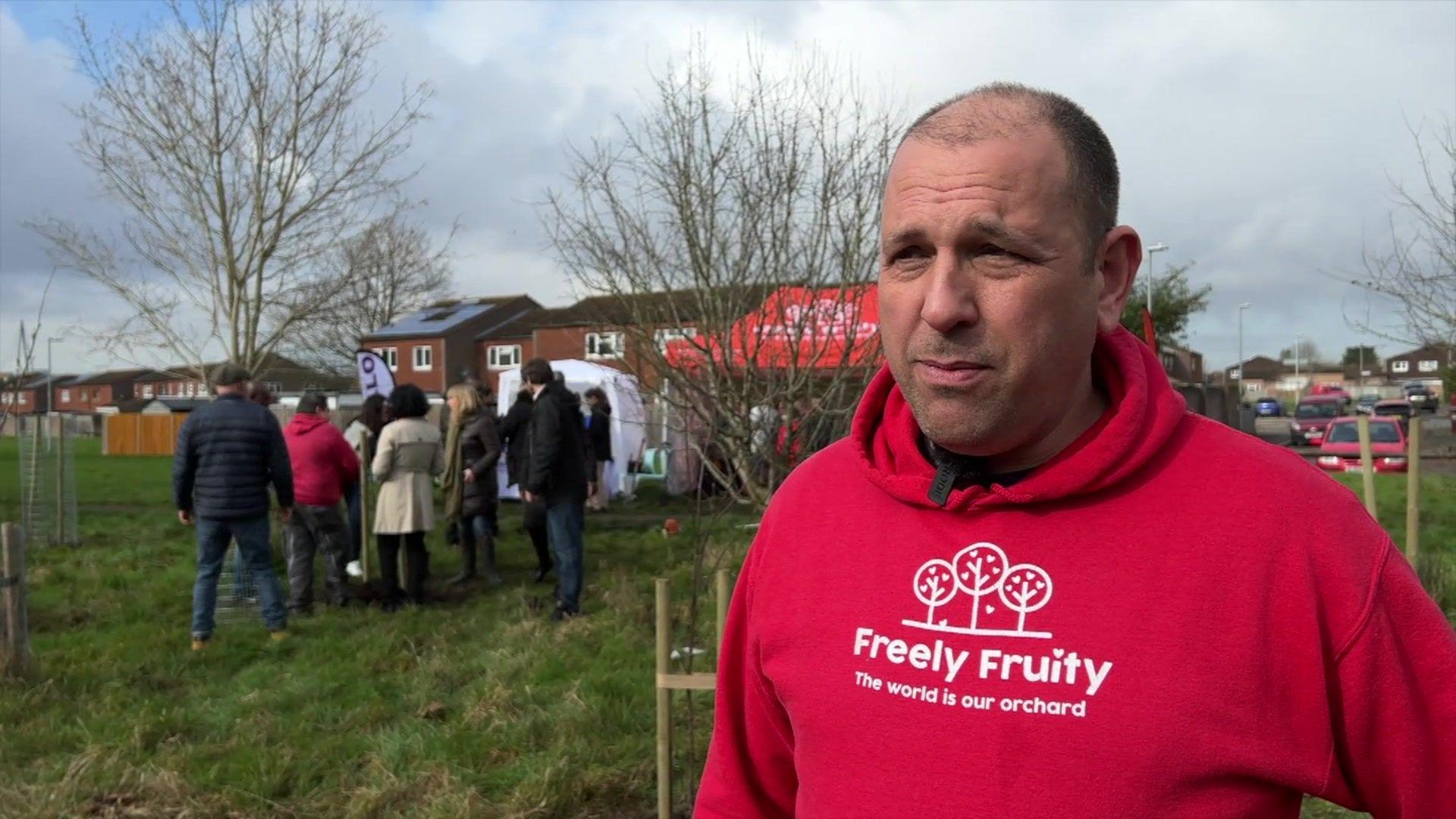 Matt Knight stands to the right facing the camera in front of a group of people who are gathered around a new tree in an open green space. He is wearing a red hoodie with the charity's logo in white on the front. He has short brown hair