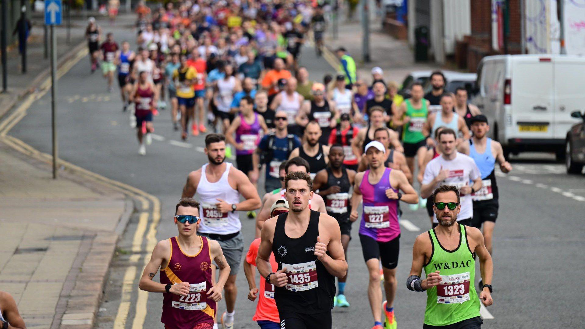 Dozens of runners race along a closed road. They have a colourful range of vests and running tops with a number of clubs including the claret and yellow of Northampton Road Runners visible.
