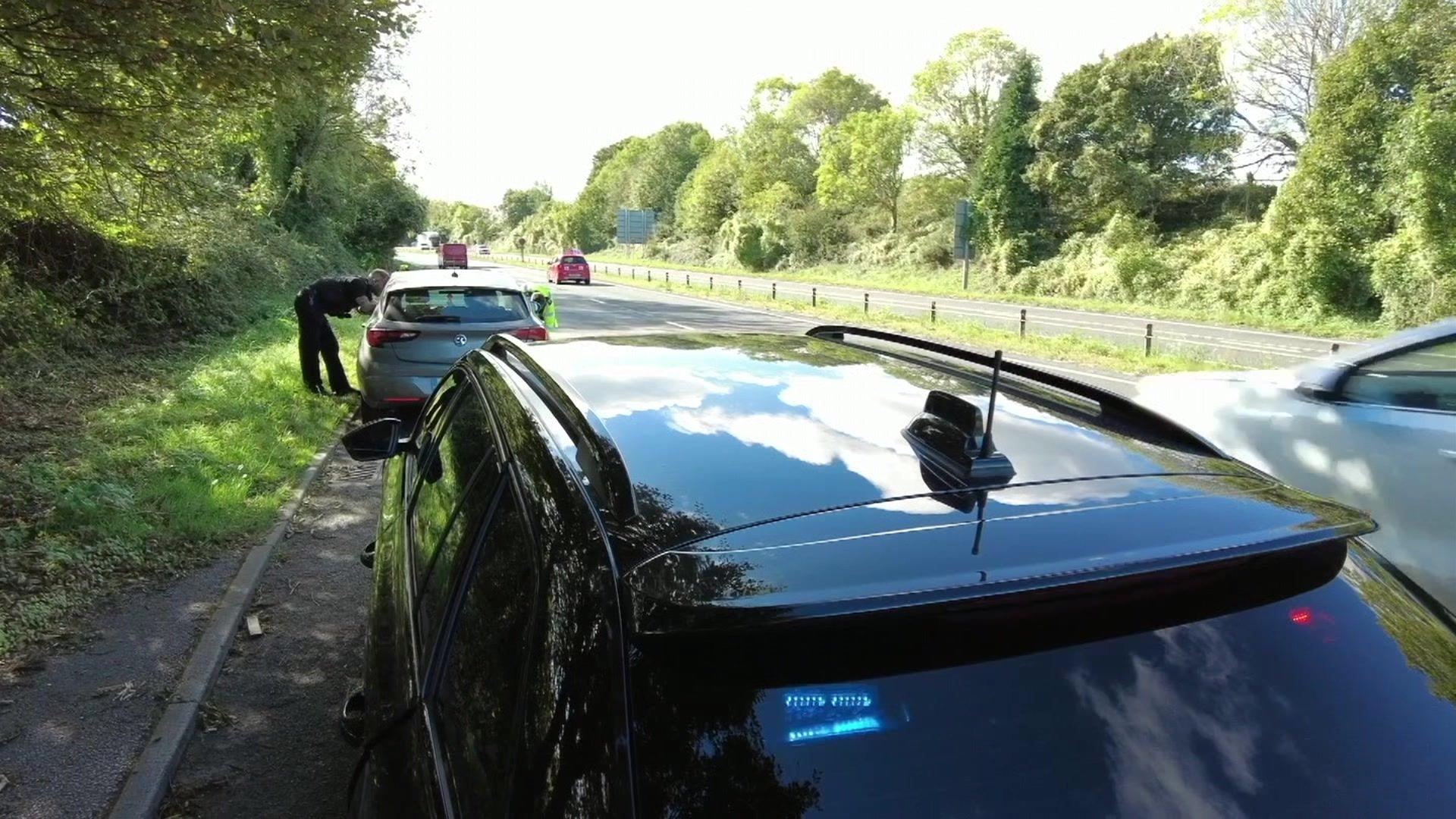 The back of an unmarked police car with blue and red lights in the rear window. There are police officers with their heads through the window of another car which is parked on the side of a motorway