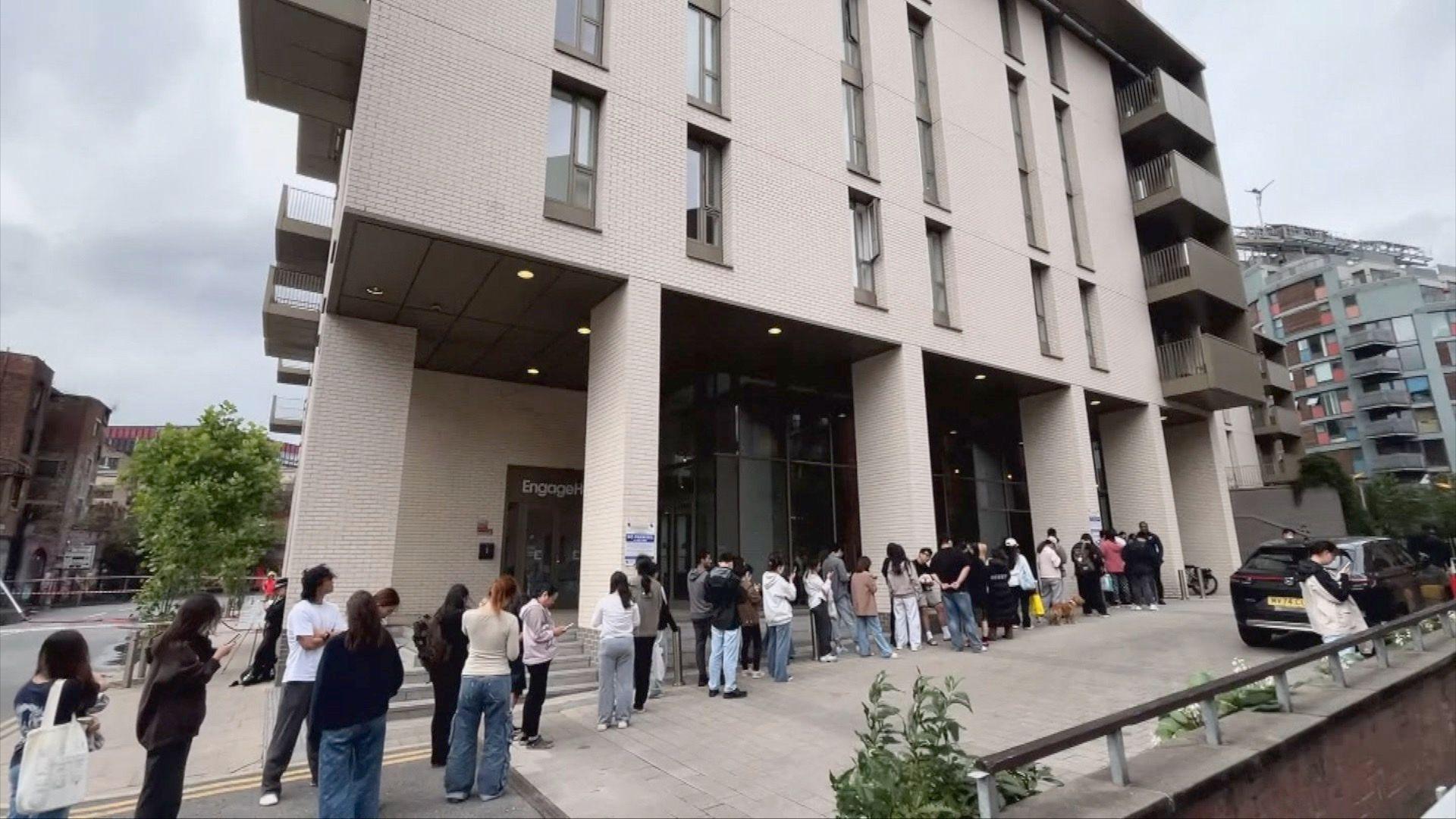 A long queue of people underneath a big cream block of flats.