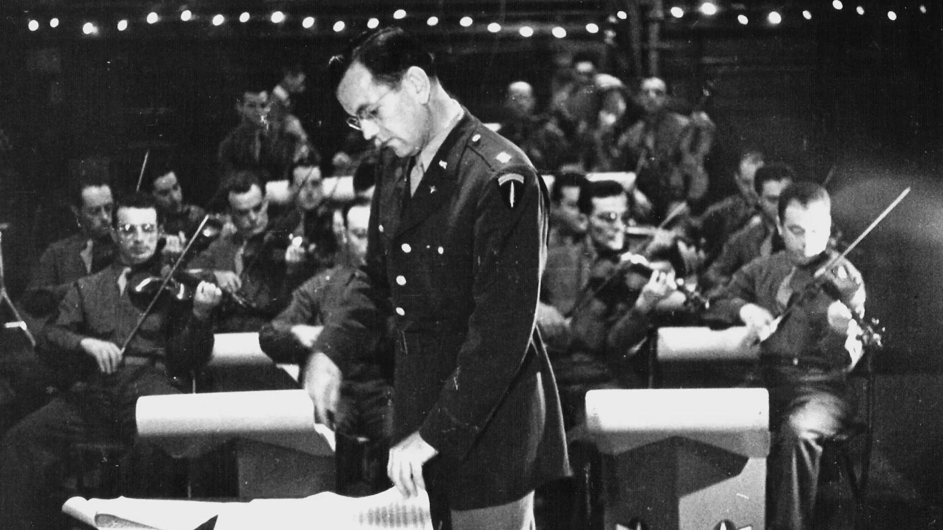 Glenn Miller seen sideways on looking down at a musical score while a large number of violinists play behind him. They are seated and all have music stands to their left or right. He wears military uniform and glasses, and has short dark hair.